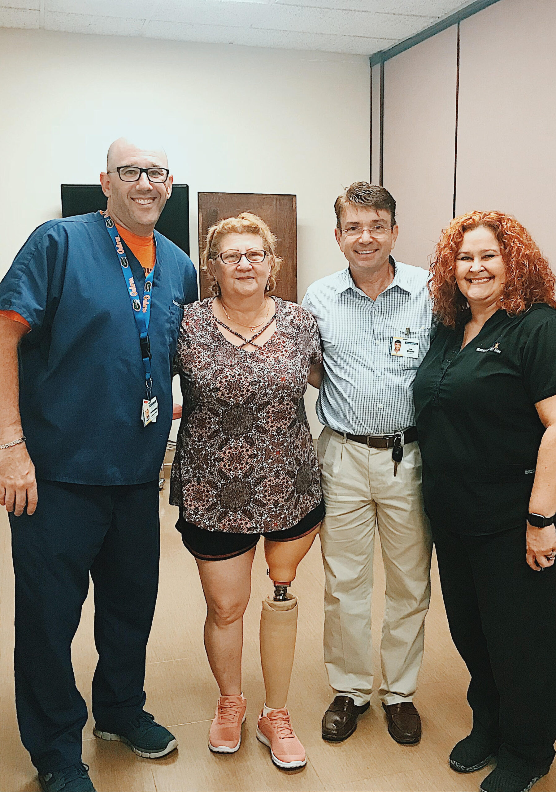 man in scrubs on far left, standing next to woman with prosthetic leg, another man on her right and a woman on his right. All are pictured smiling and posing for a picture in a clinic room.
