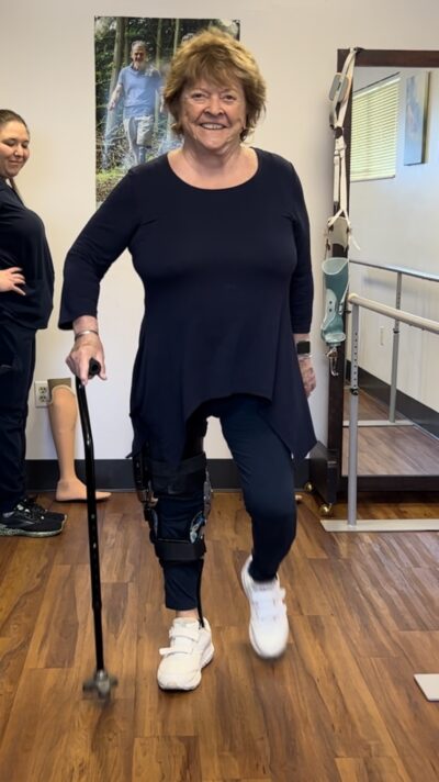 White woman with short strawberry blonde hair standing and taking a step with a cane in her right hand while wearing a leg brace.