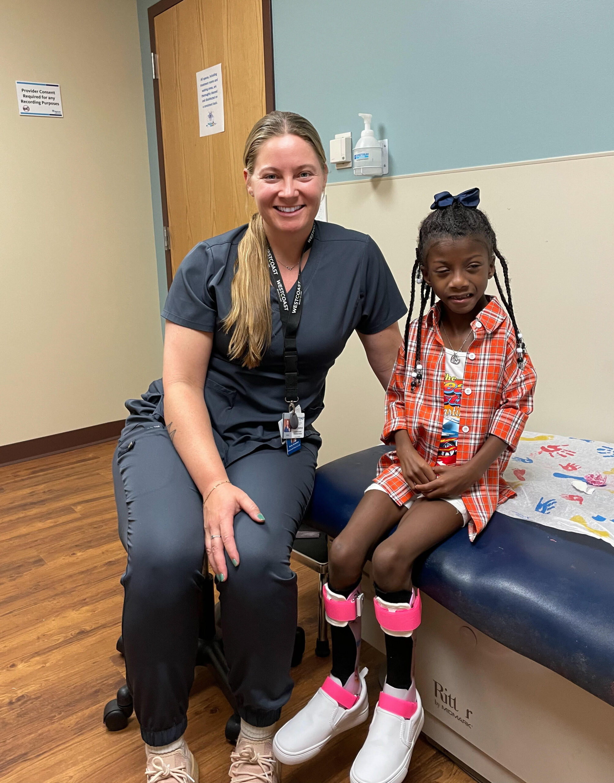 adult female with blond hair wearing scrubs sitting on medical bed with young african-american girl wearing bilateral AFO leg braces