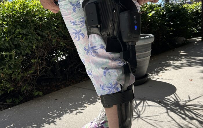 Close up of a woman's leg wearing white and floral patterned capri pants and a black Tectus microprocessor leg brace that goes from hip to foot.